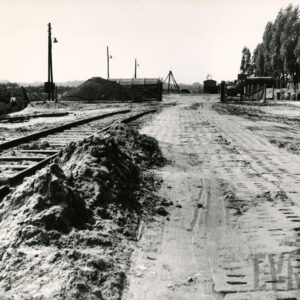 gruntowa droga wyłożona betonowymi płytami, z lewej pryzmy piachu, obok nich szyny z podkładami, z prawej barak i rząd wysokich drzew