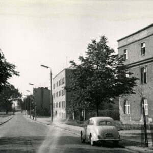 asfaltowa droga, po jej prawej stronie murowane, otynkowane, kilkukondygnacyjne budynki, na drodze samochód osobowy