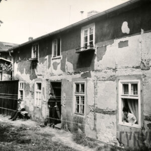 dwukondygnacyjny budynek z odpadającym tynkiem, z lewej na ławce siedzi mężczyzna, w drzwiach pośrodku stoi mężczyzna, przez okno z prawej wygląda kobieta