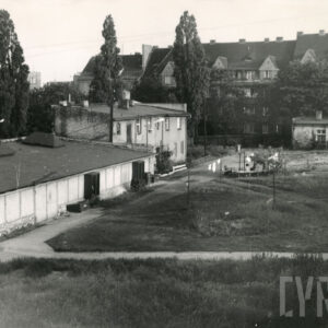 na pierwszym planie niskie murowane budynki, w głębi drzewa, za nimi kilkukondygnacyjna kamienica