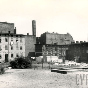 z lewej otynkowany trzykondygnacyjny budynek mieszkalny, z prawej czterokondygnacyjny budynek drewniano-ceglany w formie muru pruskiego, w głębi ściany wysokich kamienic