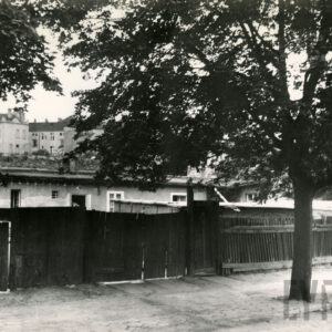 z przodu drewniany płot, przed nim, z prawej rosną dwa drzewa, za płotem widoczny parterowy otynkowany budynek, w głębi wysokie kamienice