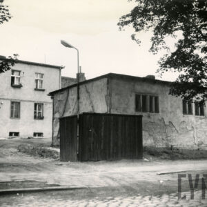 na pierwszym planie fragment drewnianego płotu z otwartą bramą, w głębi dwukondygnacyjny otynkowany, podpiwniczony budynek, z przodu z prawej parterowy budynek z odpadającym tynkiem