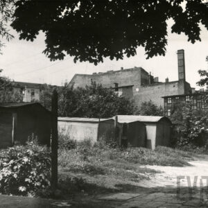 blaszane i drewniane garaże, z prawej fragment budynku drewniano-ceglanego, w głębi murowane ściany kamienic, bez okien, na jej narożniku, z prawej wysoki komin