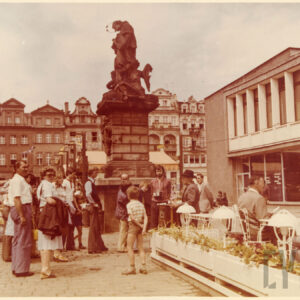 kolorowa fotografia fragmentu rynku ze stolikami i krzesłami stojącymi na wolnym powietrzu, w tle grupa ludzi stoi przed grającym kataryniarzem, w głębi pomnik świętego na wysokim postumencie