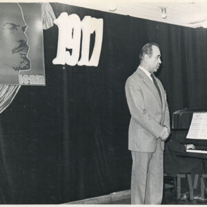 mężczyzna stoi na scenie, za nim na ciemnej kotarze plakat Lenina i rok 1917