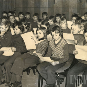 duża grupa dzieci siedzi na krzesłach, w rękach trzymają kartki papieru