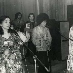 z przodu, w środku mężczyzna z mikrofonem, po jego lewej i prawej stronie dwie kobiety w kwiecistych sukniach