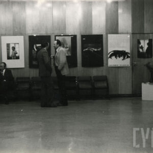 na ścianach wiszą oprawione fotografie, na sali dwie osoby siedzą na fotelach, dwóch mężczyzn stoi i rozmawia ze sobą