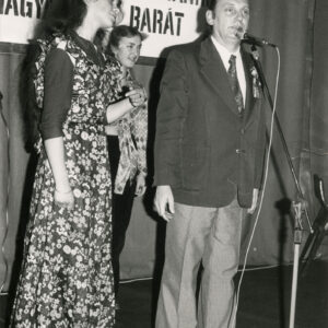 kobieta w długiej sukni w kwiaty, obok niej mężczyzna przy mikrofonie, za nimi fragment napisu w języku polskim i węgierskim