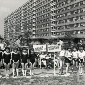 dwie grupy dzieci w strojach sportowych stoją w rzędzie, za nimi grupa dorosłych, w głębi długi, wielopiętrowy blok mieszkalny