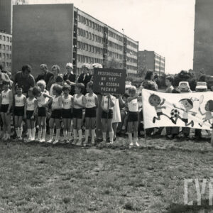 grupa dzieci w strojach gimnastycznych, za nimi dorośli, w głębi kilkupiętrowe bloki mieszkalne