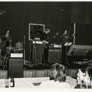 trzech mężczyzn występuje na scenie i grają na perkusji, gitarze elektrycznej i skrzypcach