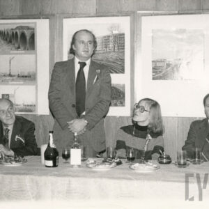 grupa ludzi siedzi przy zastawionym stole, jeden z mężczyzn stoi