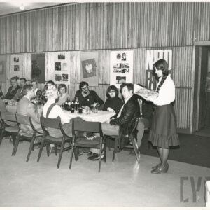 duża grupa ludzi siedzi przy zastawionym stole, z prawej stoi dziewczyna z warkoczem zwrócona w kierunku siedzących przy stole odczytuje tekst, który trzyma przed sobą