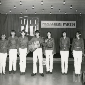 grupa dziewczyn stoi przed sceną, wszystkie z wyjątkiem jednej ubrane są w białe spodnie i ciemniejsze bluzki, pośrodku chłopak gra na gitarze