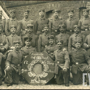 grupa mężczyzn w średnim wieku, ubranych w mundury, pozujących do zdjęcia na tle budynku z cegieł; mężczyźni pozują w trzech szeregach, czterech z nich klęczy, dwóch siedzi, reszta stoi