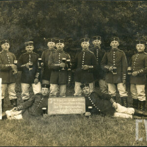 grupa młodych mężczyzn w mundurach pozująca do zdjęcia pamiątkowego, mężczyźni trzymają kufle z piwem; widoczne tabliczki okolicznościowe z napisami w języku niemieckim