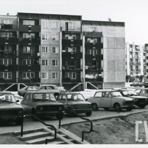 na pierwszym planie delikatna skarpa z prowadzącymi na nią schodami, na placyku zaparkowane samochody osobowe, w głębi pięciokondygnacyjne bloki mieszkalne z wielkiej płyty, z balkonami