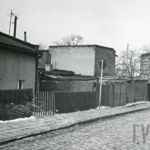 niskie budynki ceglane i murowane ogrodzone drewnianym płotem, na pierwszym planie brukowana ulica