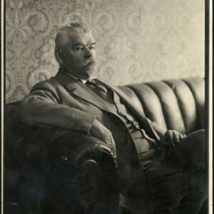 starszy, siwy mężczyzna siedzi na skórzanej kanapie, za nim na ścianie wzorzysta tapeta