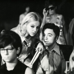 czarno-biała fotografia grupy kilku osób, w środku siedzi mężczyzna, nad nim pochyla się młoda kobieta