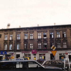 dwupiętrowa kamienica z płaskim dachem, przed kamienicą ruch uliczny