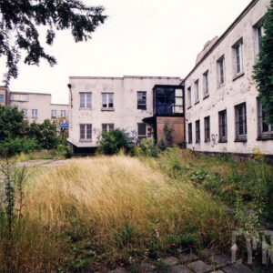 dwukondygnacyjny budynek w kształcie litery „L”, przed budynkiem wysoka trawa i krzewy