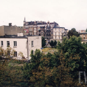 na pierwszym planie korony drzew, dalej dwukondygnacyjny budynek, w głębi kilkupiętrowe kamienice