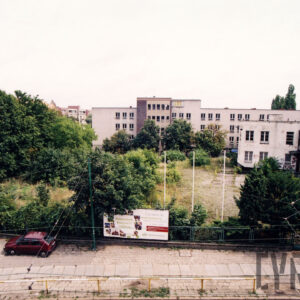 plac porośnięty drzewami, krzewami i trawą, z prawej i na wprost kilkukondygnacyjne budynki