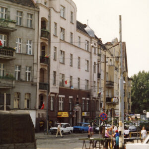 kilkupiętrowe kamienice wzdłuż brukowanej ulicy, na pierwszym planie z prawej fragment targowiska ze stoiskami