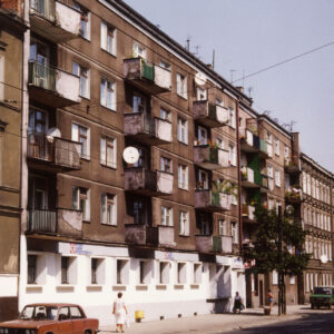 czteropiętrowy blok z balkonami, na pierwszym planie ulica z torami tramwajowymi