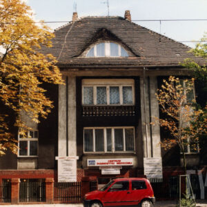 jednopiętrowy dom z czterospadowym dachem, fasada ozdobiona lizenami, przed budynkiem ogrodzenie i zaparkowany samochód