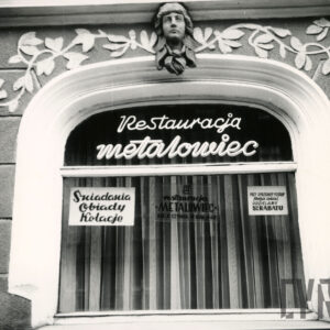 owalnie zakończone okno wystawowe z dekoracją rzeźbiarską nad nim, na szybie napis z nazwą restauracji oraz wywieszone w oknie trzy tablice: „śniadania, obiady, kolacje”, „restauracja Metalowiec czynna 9-22”, „przy sprzedaży potraw poza lokal udzielamy 5% rabatu”