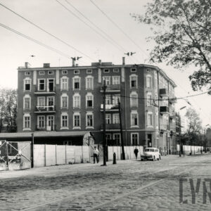 na pierwszym planie brukowana ulica z torami tramwajowymi, w głębi trzypiętrowa kamienica, wzdłuż ulicy ogrodzenie