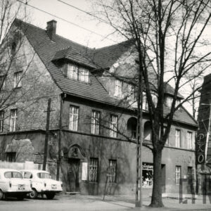 trzykondygnacyjny budynek z trójkątnym szczytem, z lewej zaparkowane samochody