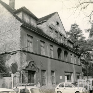 trzykondygnacyjny budynek z trójkątnym szczytem, przed nim zaparkowane samochody