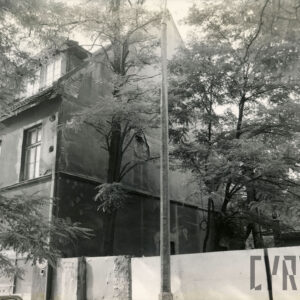 fragment budynku ze spadzistym dachem, dookoła budynku wysokie drzewa