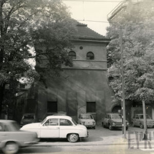 wąski budynek ze spadzistym dachem, w górnej jego części niewielki półokrągłe okna, przed budynkiem zaparkowane samochody