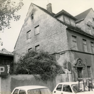 trzykondygnacyjny budynek z trójkątnym szczytem, przy nim zaparkowane samochody