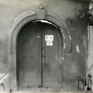 drewniane, łukowe, dwuskrzydłowe drzwi w murze, na drzwiach tabliczka ostrzegająca przed niebezpieczeństwem (elektryczność)