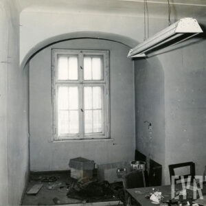 pomieszczenie z oknem, u sufity wisi lampa, w prawym dolnym rogu widoczny stół, na stole i na podłodze porozrzucane kartony i przedmioty