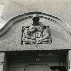 dekoracja rzeźbiarska nad drzwiami budynku przedstawiająca dwójkę, zwróconych do siebie i klęczących dzieci(?), trzymających tarczę z inicjałami JD