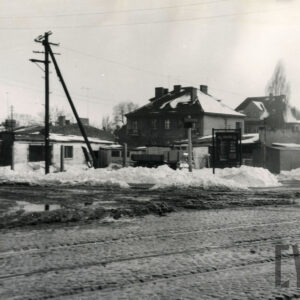 na pierwszym planie brukowana ulica z torami tramwajowymi, w głębi niskie baraki, za nimi miejskie wille, na ulicach śnieg i kałuże