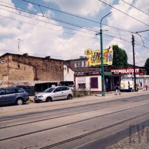 na pierwszym planie ulica częściowo asfaltowa, częściowo brukowana z torami tramwajowymi, w głębi niskie pawilony handlowe z kolorowymi szyldami, za nimi ceglane, niskie budynki