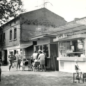 z prawej kioski handlowe, z lewej dwukondygnacyjny budynek murowany, przed kioskami kobiety z dziećmi i wózkami dziecięcymi, część stoi w kolejce