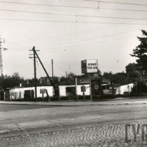 na pierwszym planie brukowana ulica z torami tramwajowymi, w głębi niska zabudowa, z lewej słupy energetyczne