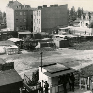 na pierwszym planie kiosk i stojący przy nim mężczyźni, w głębi garaże i szopki, za nimi kilkukondygnacyjny budynek mieszkalny