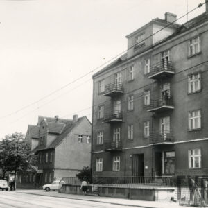 budynki mieszkalne wzdłuż ulicy, z prawej trzypiętrowy budynek z balkonami i mieszkalnym poddaszem, za nim jednopiętrowy budynek ze spadzistym dachem i mieszkalnym poddaszem 