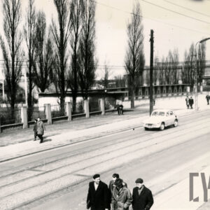 częściowo brukowana, a częściowo asfaltowa ulica z torami tramwajowymi, ogrodzenie z małymi murowanymi kioskami, za ogrodzeniem wysokie drzewa